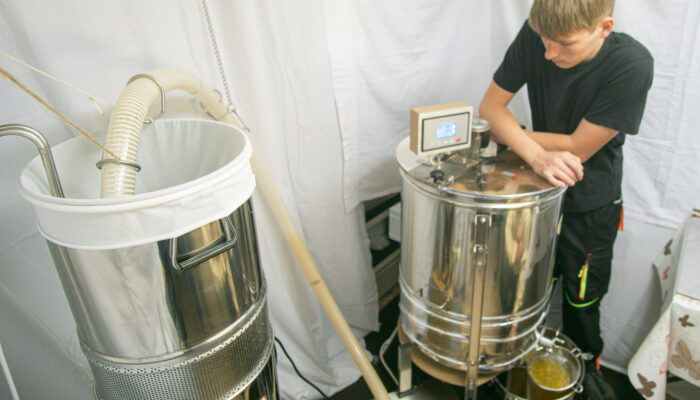 Jungimker Jannik steht neben der Honigschleuder, die mit einem Schlauch an das Honigsieb angeschlossen ist. Jannik überwacht den Vorgang und passt auf, dass alles richtig funktioniert.